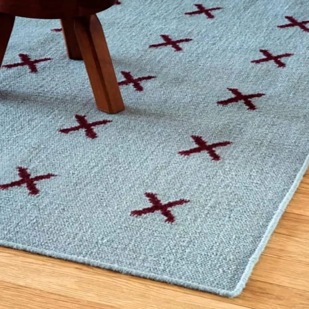Scandinavian handwoven flatweave wool rug with a minimalist burgundy cross geometric pattern on a light blue background, handmade Nordic area rug for modern living room and bedroom.
